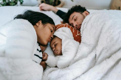 A family in bed, with a sleeping father, a mother kissing her newborn baby, and the baby resting comfortably between them.